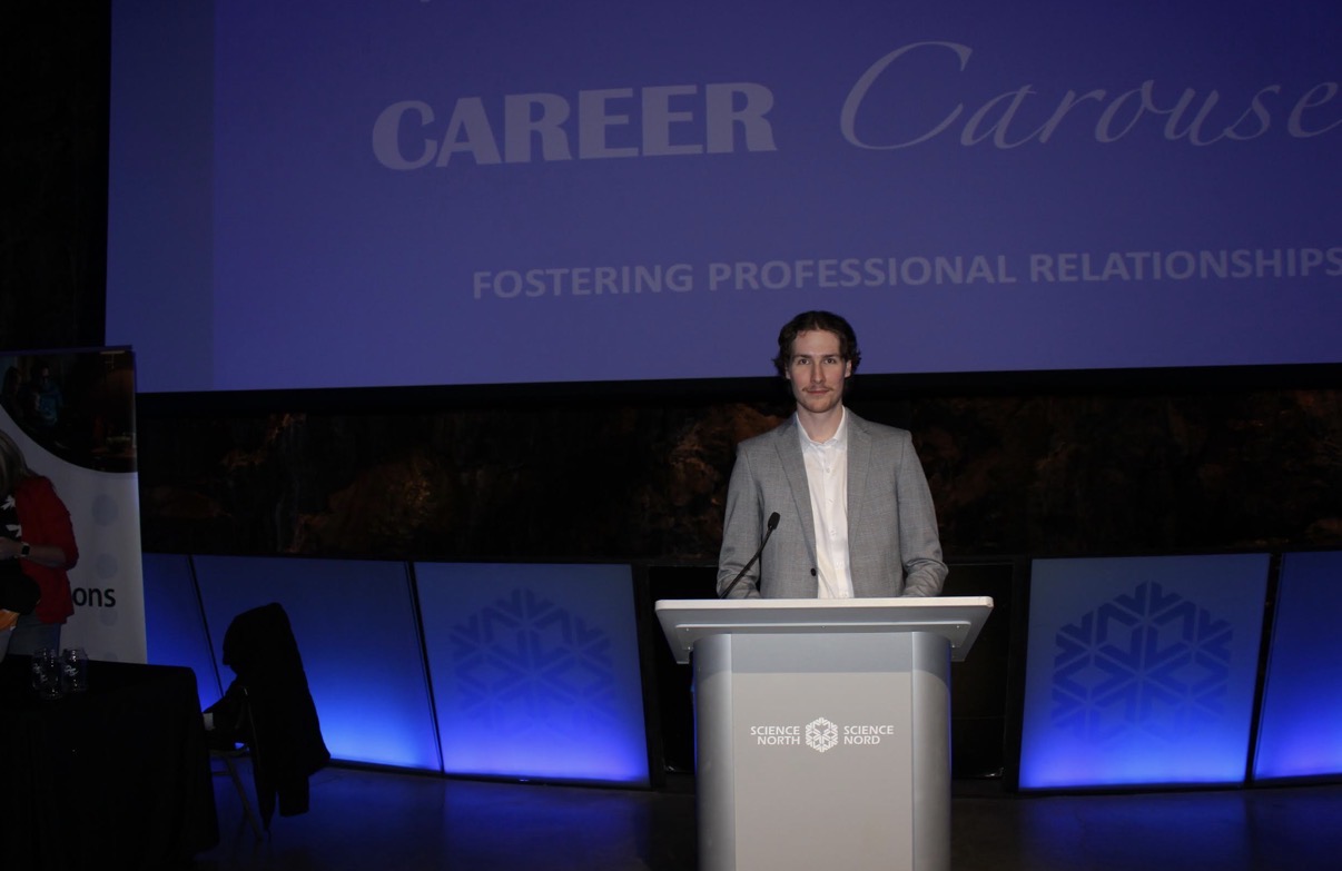 A photograph of Paul Petitclerc standing confidently behind a silver podium inside a dimly lit auditorium. He is wearing a white button-down shirt paired with a grey blazer. Behind him, a massive projector screen displays a presentation slide reading "CAREER Carousel" and "FOSTERING PROFESSIONAL RELATIONSHIPS." The podium features a prominent logo for Science North.