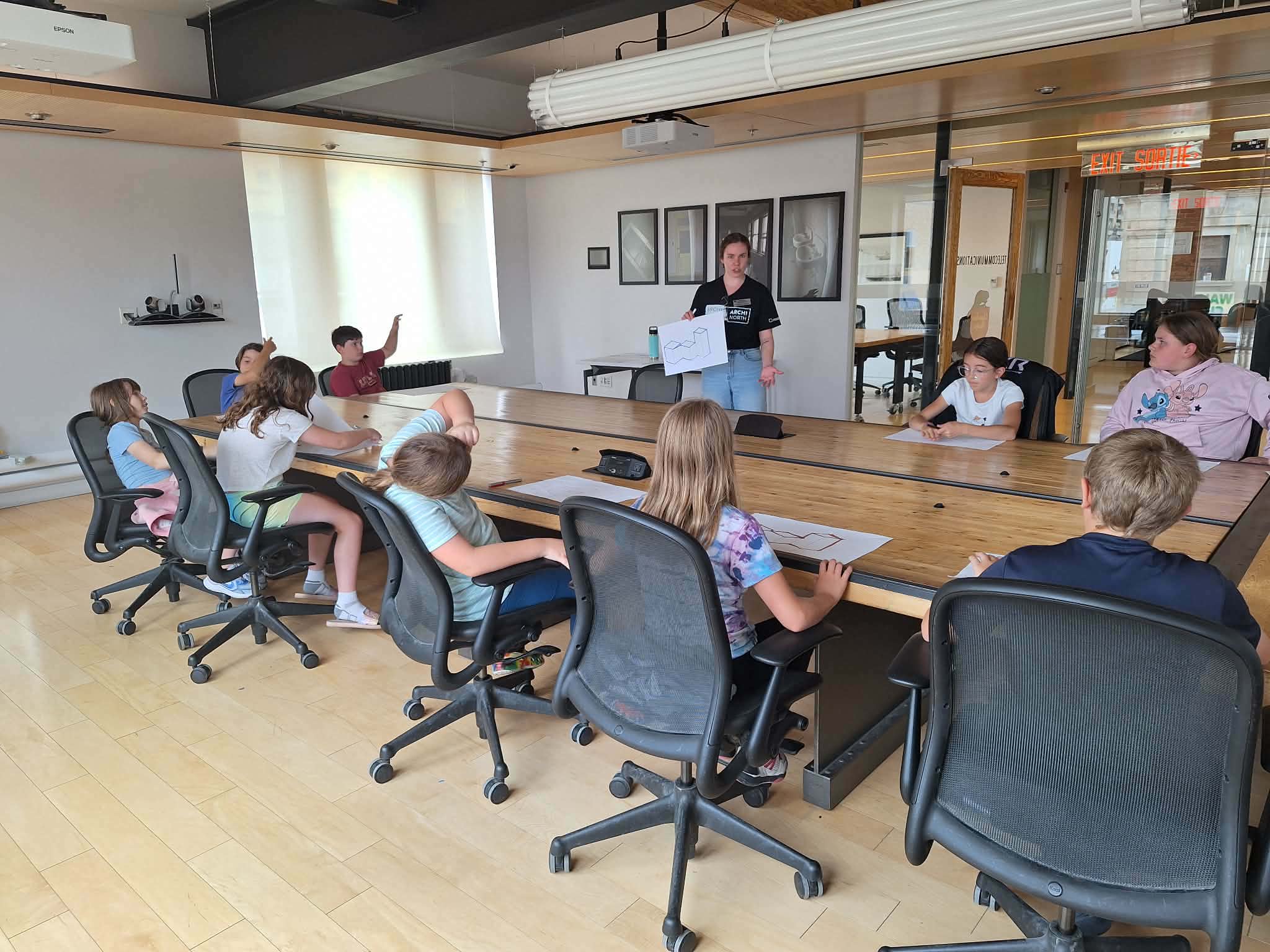 An instructor stands at the head of a large conference table, holding up a paper design proposal while youth campers listen and raise their hands to participate in the design lesson.