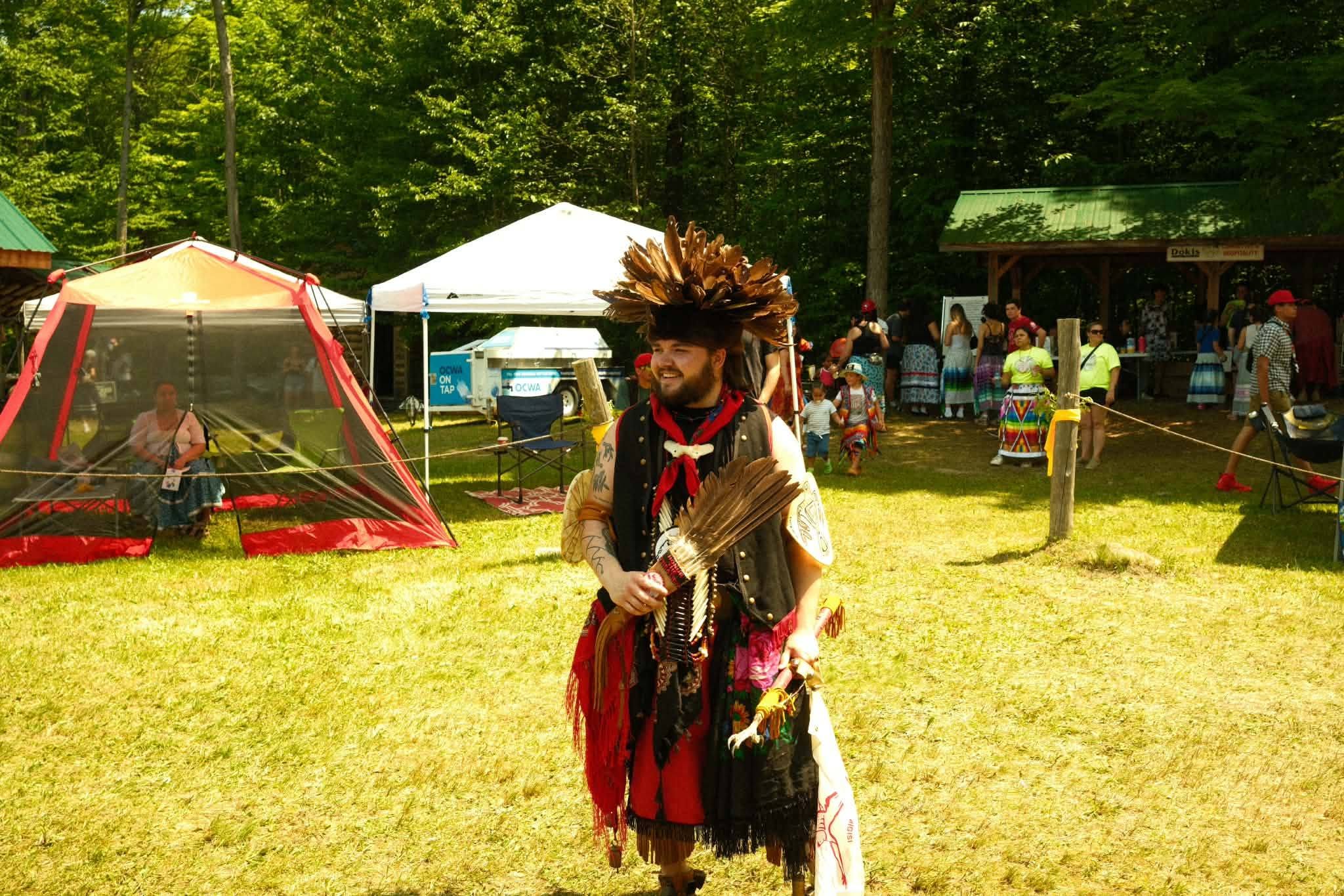 Header: Connor Lafortune wearing traditional Anishinaabe regalia and a feather hat while holding feather fans, smiling outdoors at an Indigenous powwow event.