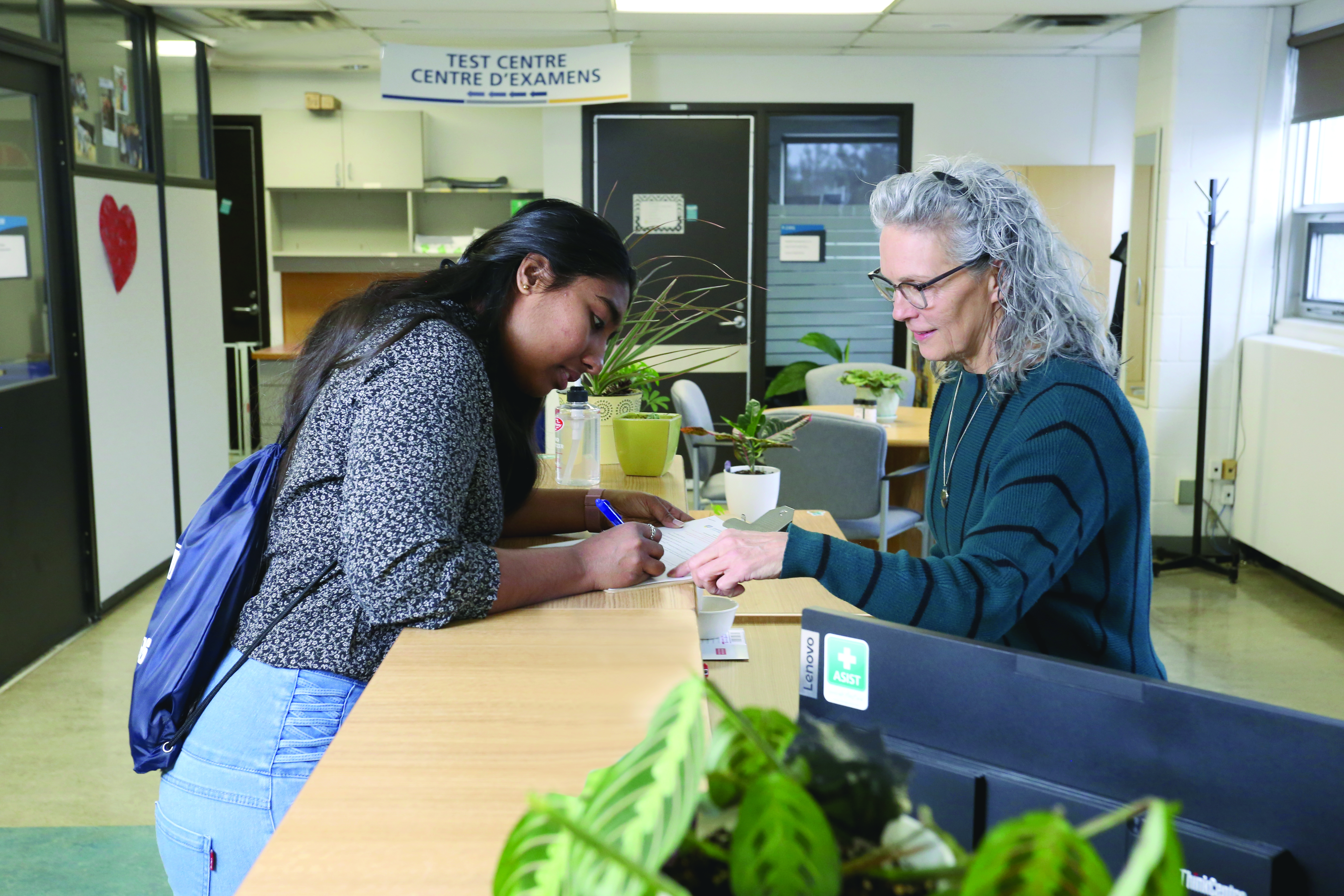A student leaning over a front desk, filling out a form with a pen while a staff member (intake coordinator) points to a section on the paper.