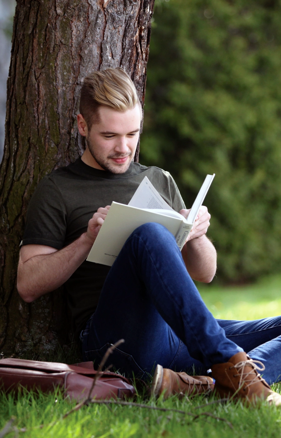 A Laurentian University student sitting outside on campus under a tree and reading a book.